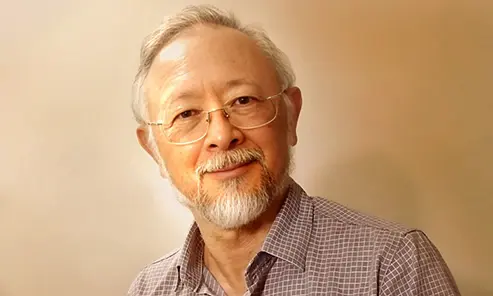 Roberto Otsu, um senhor de traços orientais, usando óculos de aro, cabelos e cavanhaque grisalhos, sorri e olha para a câmera.